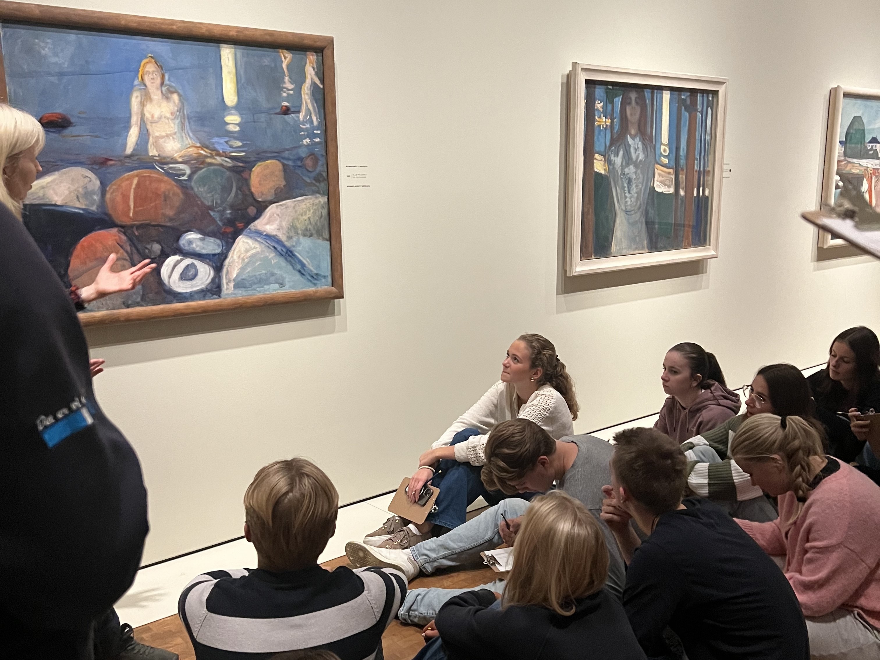 en gruppe mennesker som sitter i et rom og ser på kunst på veggen